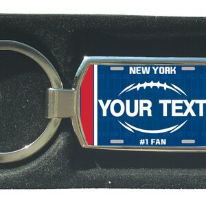 Könnte beinhalten: Ein silberner Schlüsselanhänger mit einem blauen und roten Nummernschild-Design. Das Nummernschild zeigt "NEW YORK" und hat eine Fußballgrafik. Der Text "YOUR TEXT" ist auf dem Nummernschild gedruckt. Der untere Teil des Nummernschilds lautet "#1 FAN".