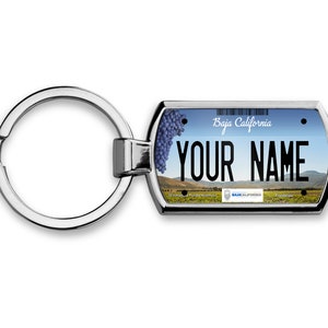 Może przedstawiać: Srebrny brelok do kluczy z prostokątnym znacznikiem przedstawiającym projekt tablicy rejestracyjnej Baja California. Znacznik ma niebieskie niebo, zielone wzgórza i grono winogron. Tekst "Baja California" i "YOUR NAME" jest nadrukowany na znaczniku czarnym kolorem.