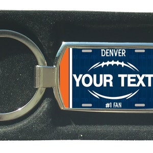Könnte beinhalten: Ein silberner Schlüsselanhänger mit einem blauen und orangenen Nummernschild-Design. Das Nummernschild zeigt "DENVER" und "#1 FAN". Der Text "YOUR TEXT" befindet sich in der Mitte des Nummernschilds.