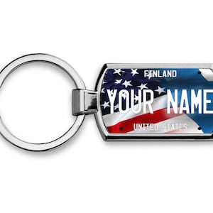 May include: A silver keychain with a rectangular tag featuring a waving American flag design. The tag has the text "FINLAND" and "UNITED STATES" at the top and bottom, with "YOUR NAME" in the center.