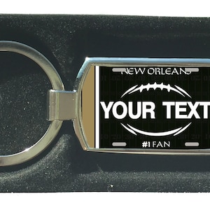 Könnte beinhalten: Ein silberner Schlüsselanhänger mit einem rechteckigen Anhänger in Schwarz und Gold. Der Anhänger zeigt ein Fußball-Design mit dem Text "NEW ORLEANS" und "YOUR TEXT" in Weiß. Der Anhänger trägt auch den Text "#1 FAN" in Weiß.