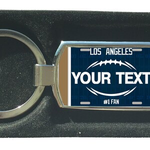 Könnte beinhalten: Ein silberner Schlüsselanhänger mit einem blauen und braunen Design zum Thema Fußball. Der Schlüsselanhänger trägt den Text "LOS ANGELES" und "YOUR TEXT" mit den Worten "#1 FAN" am unteren Rand.