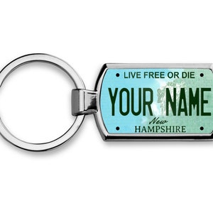 May include: A silver-colored metal keychain with a rectangular tag. The tag features a light blue and green background with the text "LIVE FREE OR DIE" and "YOUR NAME" in dark green. The words "New Hampshire" are also visible.