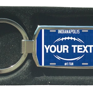 Könnte beinhalten: Ein silberner Schlüsselanhänger mit einem blauen und weißen Nummernschild-Design. Das Nummernschild zeigt "Indianapolis" und "#1 Fan". Der Schlüsselanhänger hat außerdem eine Fußballgrafik und den Text "YOUR TEXT".