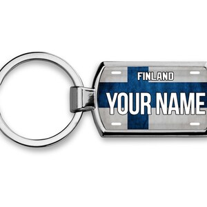 May include: A silver keychain with a blue and white Finnish flag design. The keychain has a metal tag with the text "FINLAND" and "YOUR NAME" printed on it.