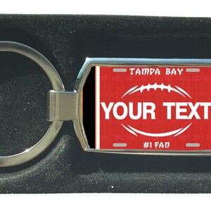 Könnte beinhalten: Ein silberner Schlüsselanhänger mit einem roten und schwarzen Nummernschild-Design. Das Nummernschild zeigt "Tampa Bay" und "#1 Fan". Der Schlüsselanhänger hat eine Fußballgrafik und den Text "YOUR TEXT".