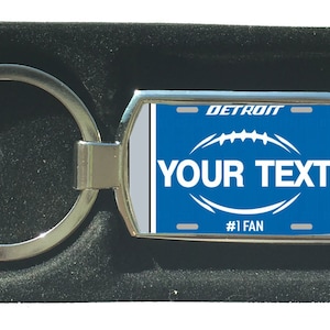 Könnte beinhalten: Ein silberner Schlüsselanhänger mit einem blauen und weißen Nummernschild-Design. Das Nummernschild zeigt "DETROIT" und "#1 FAN". Das Nummernschild hat eine Fußballgrafik und den Text "YOUR TEXT".