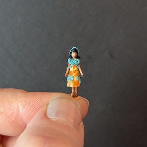 May include: A tiny doll, held between fingers, is wearing an orange dress with blue details and a matching head covering. The doll's small size and intricate clothing suggest it is handmade, possibly for a dollhouse or miniature collection.