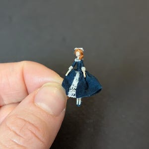 May include: A miniature doll dressed in a dark blue dress with white lace trim and a matching white bonnet. The doll has red hair and is held between fingers against a dark background. The doll is a handmade craft item.