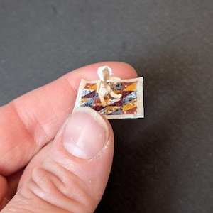 May include: A miniature figurine of a baby wearing a white bonnet and diaper, sitting on a small rectangular quilt featuring a multicolored geometric triangle pattern in shades of blue, yellow, red, and white with a cream-colored border.