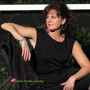 May include: A woman wearing a black sleeveless dress with a turquoise bracelet and earrings. She is sitting on a chair with a brown throw blanket. The text "RoSan Custom Jewelry" is visible in the bottom left corner of the image.