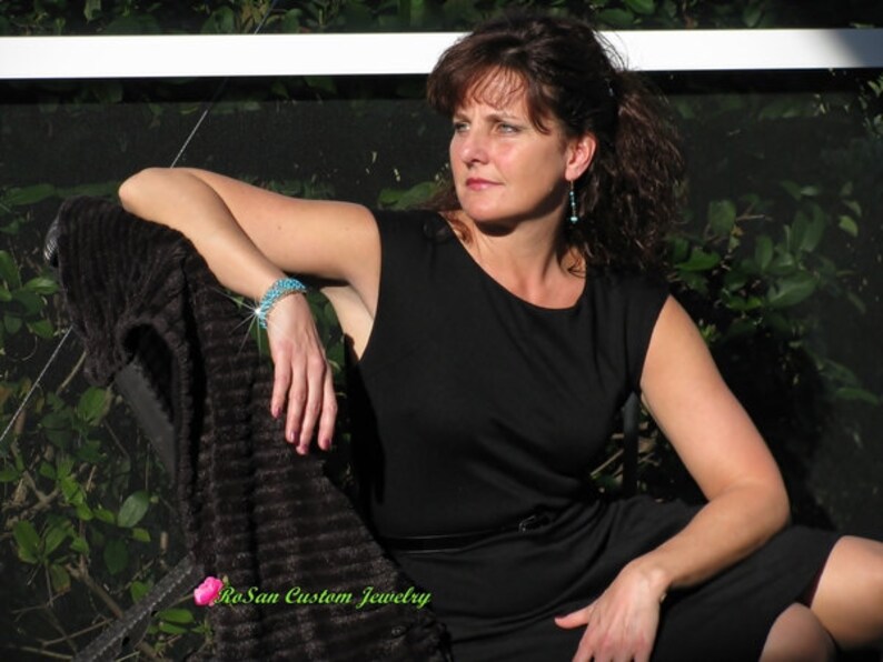 May include: A woman wearing a black sleeveless dress with a turquoise bracelet and earrings. She is sitting on a chair with a brown throw blanket. The text "RoSan Custom Jewelry" is visible in the bottom left corner of the image.