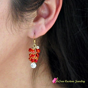 May include: Gold dangle earrings with red and clear crystal beads. The earrings are hanging from a person's ear.