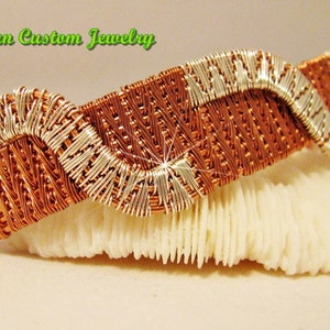 May include: A woven copper and silver wire bracelet with a wavy design. The bracelet is on a white coral-like surface.