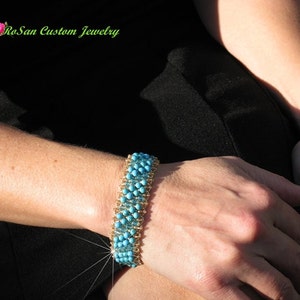 May include: A turquoise and gold beaded bracelet with a woven design. The bracelet is on a person's wrist.