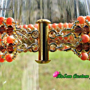 May include: A gold chain bracelet with orange and brown beads. The bracelet is made with Swarovski elements and has a clasp closure. The bracelet is displayed on a glass surface.