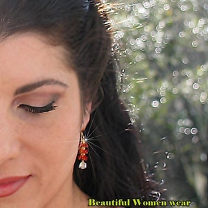 May include: A close-up of a woman's face with long dark hair. She is wearing a pink shirt and a pair of dangling earrings with red and orange beads. The text "Beautiful Women wear RoSan Custom Jewelry" is visible at the bottom of the image.
