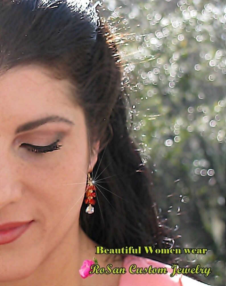 May include: A close-up of a woman's face with long dark hair. She is wearing a pink shirt and a pair of dangling earrings with red and orange beads. The text "Beautiful Women wear RoSan Custom Jewelry" is visible at the bottom of the image.