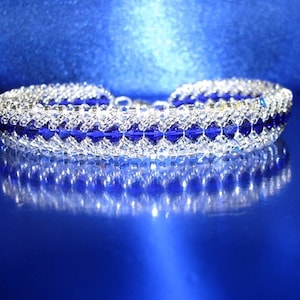 May include: A silver and blue beaded bracelet with a geometric pattern. The bracelet is reflected in a blue surface.