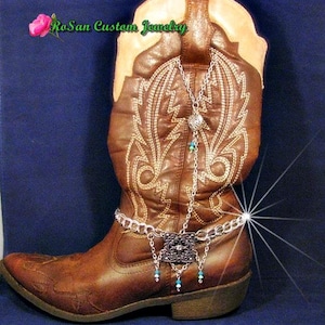 May include: A brown leather cowboy boot with a silver chain and charm dangling from the top. The chain has a silver square charm with a decorative design and turquoise beads hanging from it. The boot has a white stitching design on the shaft.