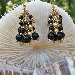 May include: Gold dangle earrings with black and gold beads. The earrings are hanging from a white coral-like surface.