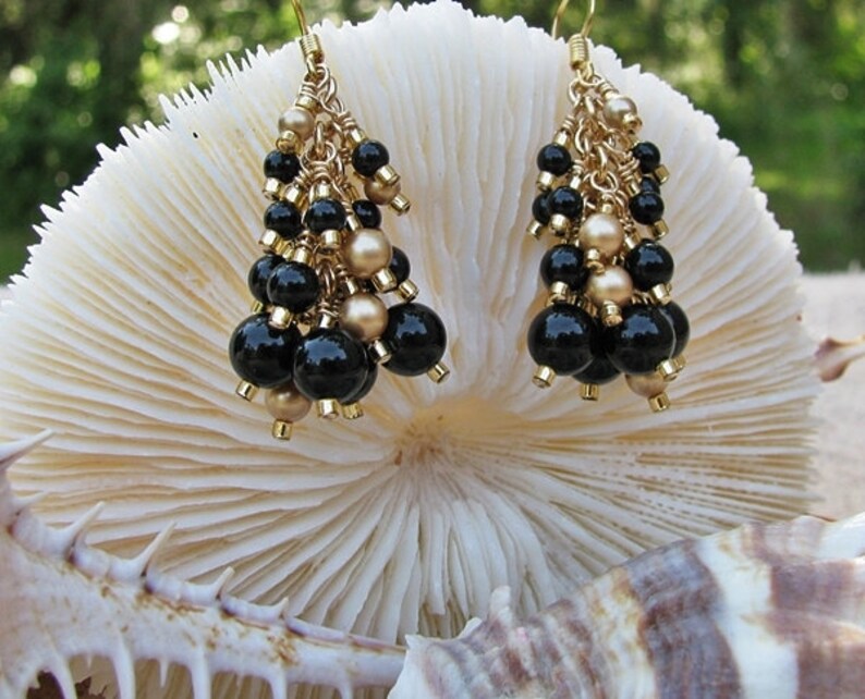 May include: Gold dangle earrings with black and gold beads. The earrings are hanging from a white coral-like surface.