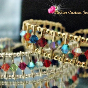 May include: Two gold wire bracelets with colourful crystal beads. The bracelets are stacked on top of each other. The top bracelet has red, blue, and iridescent beads. The bottom bracelet has red, blue, green, and iridescent beads.