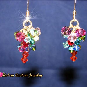 May include: A pair of gold-coloured dangle earrings. Each earring features a cluster of colourful, faceted beads in shades of pink, green, blue, and red. The earrings are suspended from gold-coloured hooks.