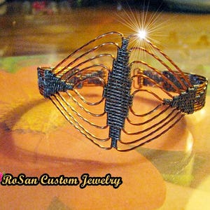 May include: A handcrafted wire bracelet with a distinctive design. The bracelet combines copper-toned wires with dark grey woven sections. The design incorporates a central vertical feature and radiating curved wires. The text "RoSan Custom Jewelry" is visible.