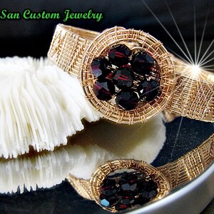 May include: Gold wire wrapped bracelet with a circular design featuring a cluster of dark red crystals. The bracelet is on a white coral-like surface.