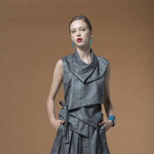 May include: A grey sleeveless dress with a draped neckline and a pleated skirt. The dress has a blue bracelet on the left wrist.