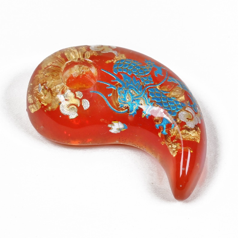 Puede incluir: Un amuleto de vidrio rojo con un dise&ntilde;o de drag&oacute;n azul y dorado. El amuleto tiene forma de media luna y tiene un agujero en la parte superior para colgarlo.