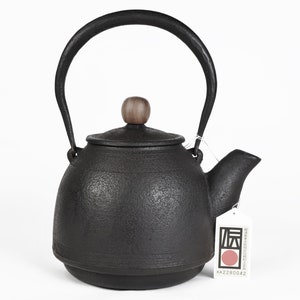 Puede incluir: Tetera de hierro fundido negro con un pomo de madera en la tapa. La tetera tiene una superficie texturizada y un pico en el lateral. La tetera está sobre una superficie blanca. La etiqueta de la tetera dice "原" en caracteres japoneses y "KA2280042".