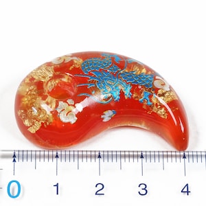 Puede incluir: Un amuleto de vidrio rojo con un dise&ntilde;o de drag&oacute;n azul y dorado. El amuleto tiene forma de media luna y mide aproximadamente 9 cm de largo.