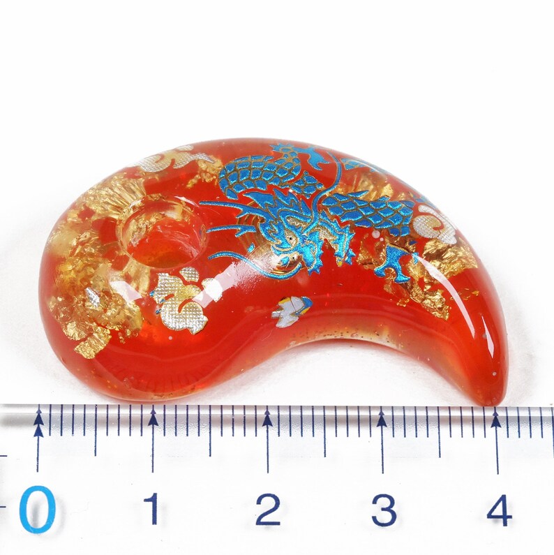 Puede incluir: Un amuleto de vidrio rojo con un dise&ntilde;o de drag&oacute;n azul y dorado. El amuleto tiene forma de media luna y mide aproximadamente 9 cm de largo.