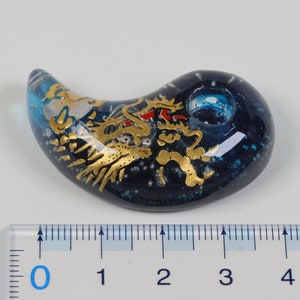 May include: A blue glass pendant with a gold dragon design. The pendant is shaped like a crescent moon and has a small hole at the top for hanging.