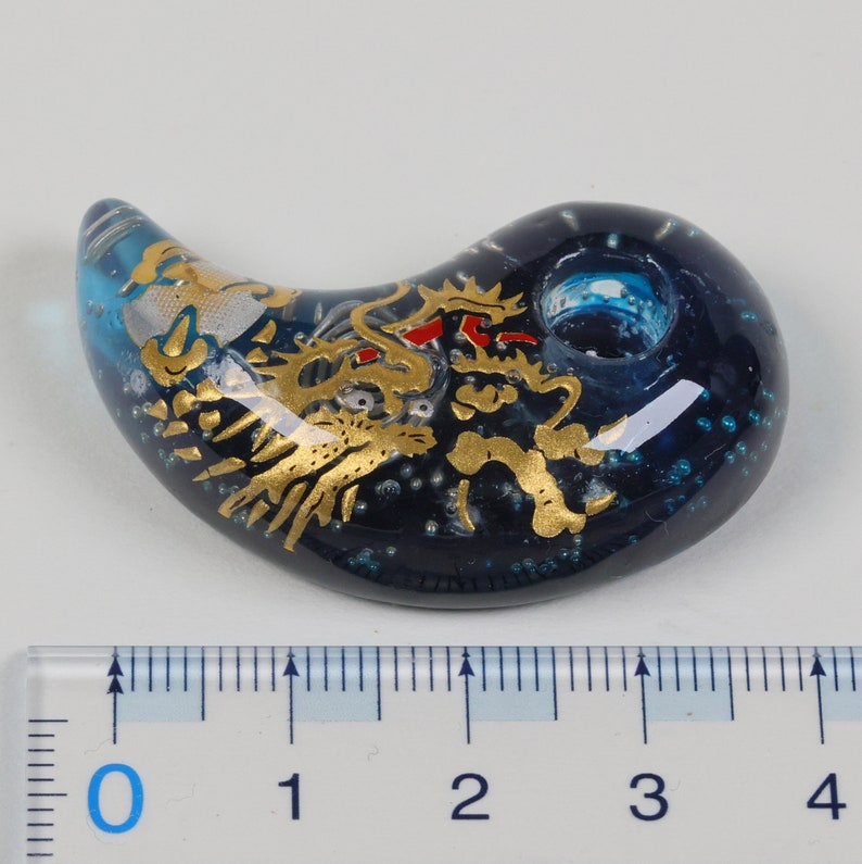 May include: A blue glass pendant with a gold dragon design. The pendant is shaped like a crescent moon and has a small hole at the top for hanging.
