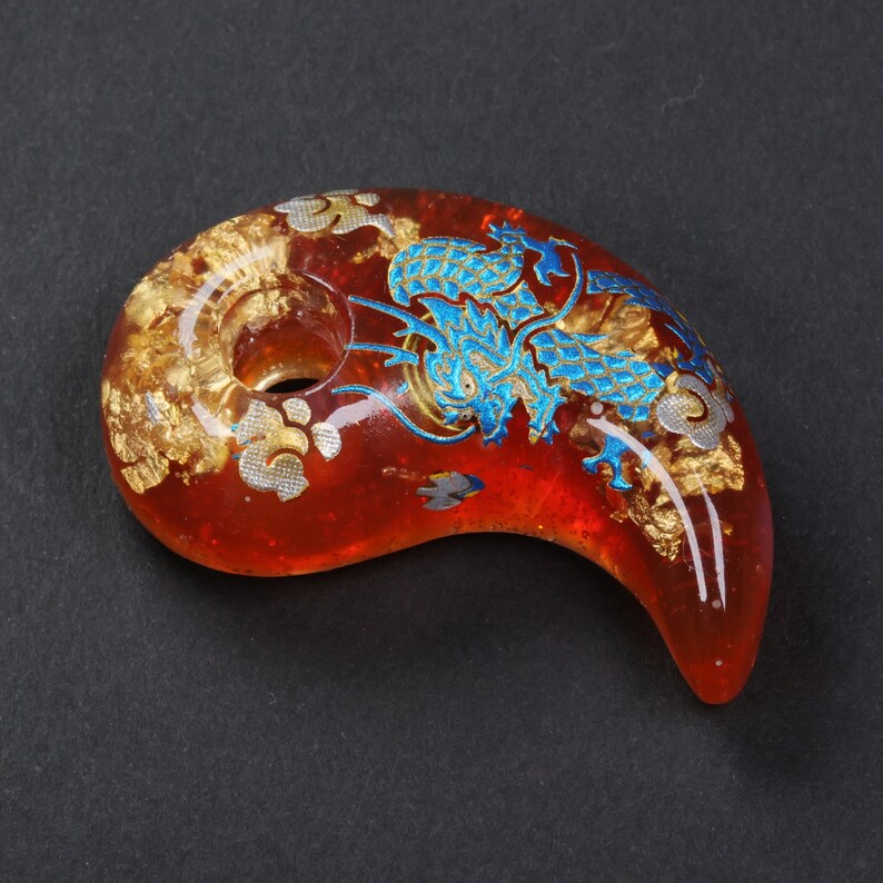Puede incluir: Un colgante de vidrio rojo con un dise&ntilde;o de drag&oacute;n azul y detalles dorados. El colgante tiene un agujero en la parte superior para un cord&oacute;n o cadena.