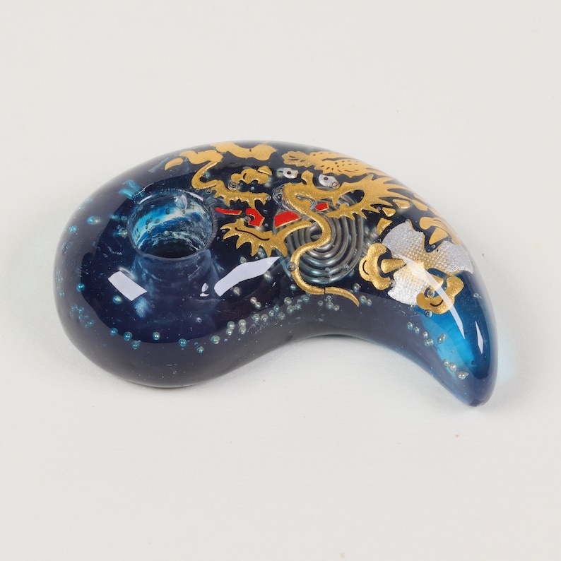 May include: A blue glass pendant with a gold dragon design. The pendant is shaped like a crescent moon and has a small hole at the top for hanging.