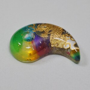 May include: A green, yellow, orange, red, purple, and blue resin pendant with gold and white accents. The pendant is shaped like a crescent moon and features a dragon design.