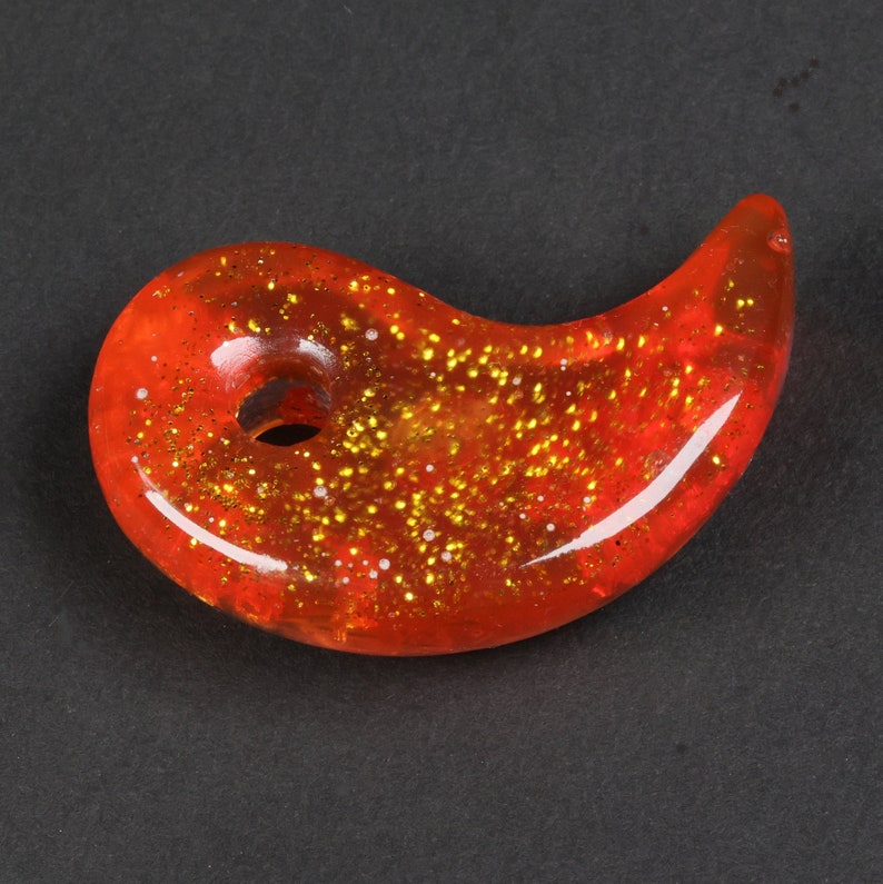 Puede incluir: Un amuleto rojo y dorado con purpurina en forma de s&iacute;mbolo yin yang. El amuleto tiene un agujero en el centro para sujetarlo a un collar o pulsera.