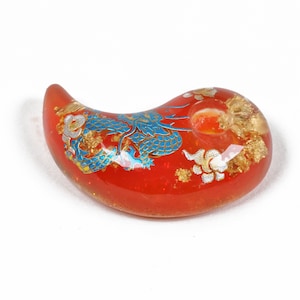 Puede incluir: Un amuleto de vidrio rojo con un dise&ntilde;o de drag&oacute;n azul y dorado. El amuleto tiene forma de media luna y tiene un agujero en la parte superior para colgarlo.