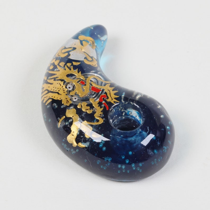 May include: A blue glass pendant with a gold dragon design and red accents. The pendant is shaped like a crescent moon and has a hole in the center for a cord.