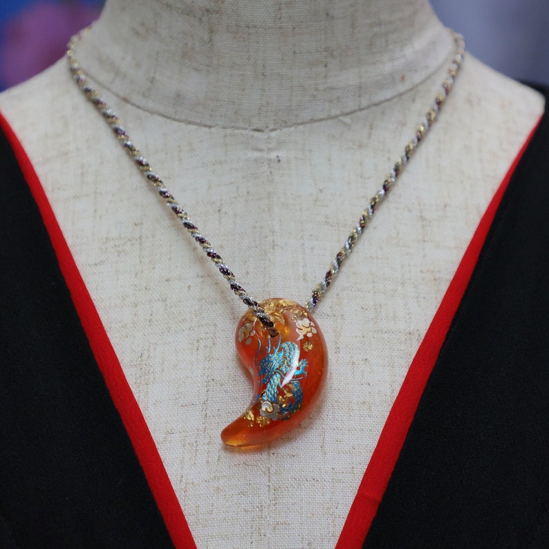Puede incluir: Un collar de cadena plateada con un gran colgante naranja con un dise&ntilde;o de drag&oacute;n azul y dorado.