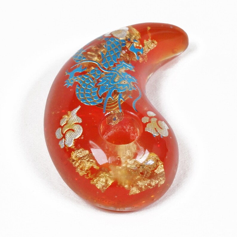 Puede incluir: Pipa de vidrio roja con un dise&ntilde;o de drag&oacute;n azul y dorado. La pipa tiene forma de media luna y tiene un agujero en el centro.