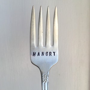 May include: A silver fork with four tines. The word "HANGRY" is stamped in black letters on the fork's handle. The fork has a decorative pattern on the handle.
