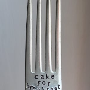 May include: Close-up of a silver fork with four tines. The words "cake for breakfast" are stamped on the handle. The fork is a kitchen utensil, perfect for eating cake.