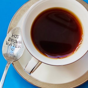 May include: A white teacup and saucer filled with dark brown liquid, likely coffee, sits on a blue surface. A silver spoon with the words "HOT BROWN WATER" and a coffee cup graphic rests beside the cup.
