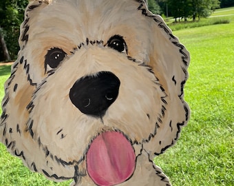 May include: A hand-painted wooden cutout of a dog's face. The dog has a light brown coat, black eyes, a black nose, and a pink tongue sticking out. The cutout is hanging from a string.
