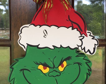 Puede incluir: Una cara verde de Grinch con ojos rojos y un gorro de Papá Noel blanco y rojo con un lazo dorado y rojo. La cara de Grinch es una decoración navideña.